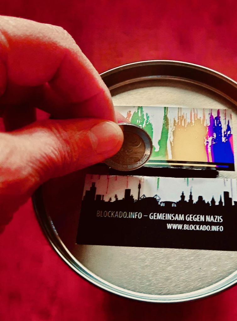 Roter Hintergrund. Finger stecken eine Münze in den Schlitz
einer Spendenbox, die einen mehrfarbigen BlockaDO-Aufkleber
auf dem Deckel hat.